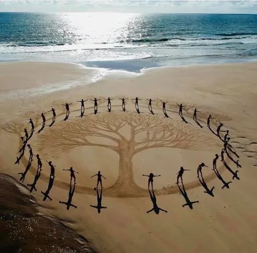 Tree of life sand art surrounded by people standing in a circle on a beach — symbolising community, connection, and growth